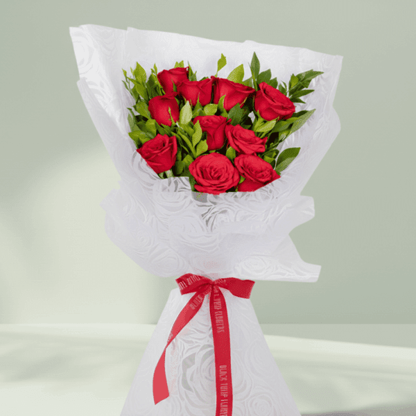 Vibrant red roses with fresh green leaves, elegantly wrapped in white non-woven paper and tied with a bright red satin ribbon.